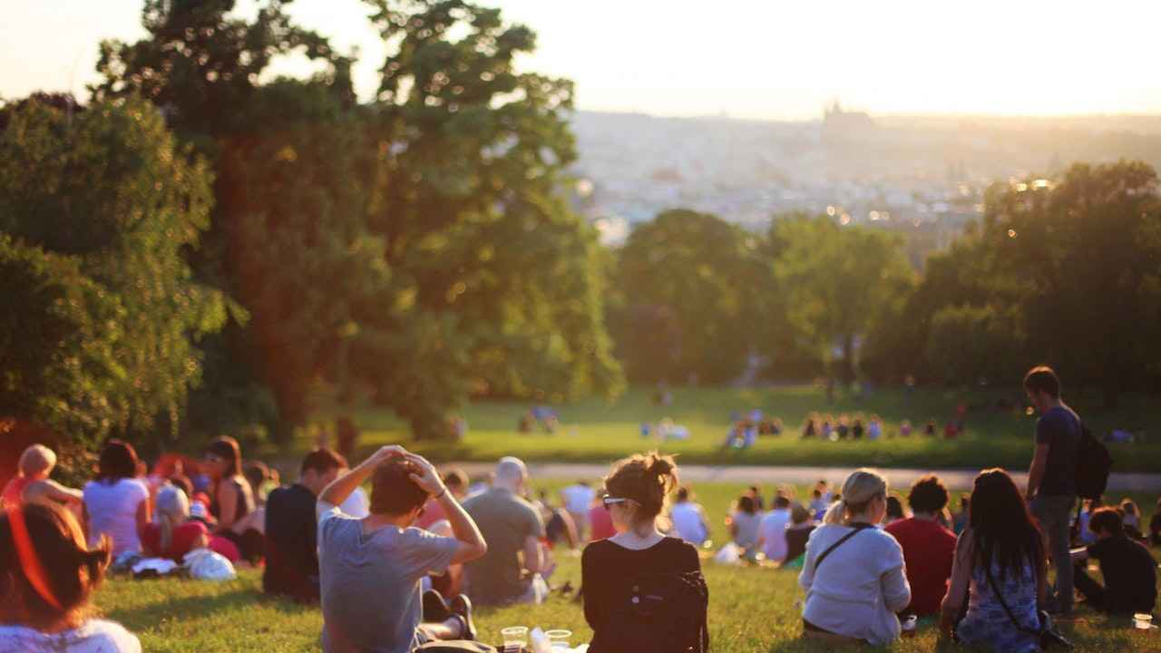 Menschen sitzen auf einer großen Wiese in einem Park und schauen in den Sonnenuntergang.